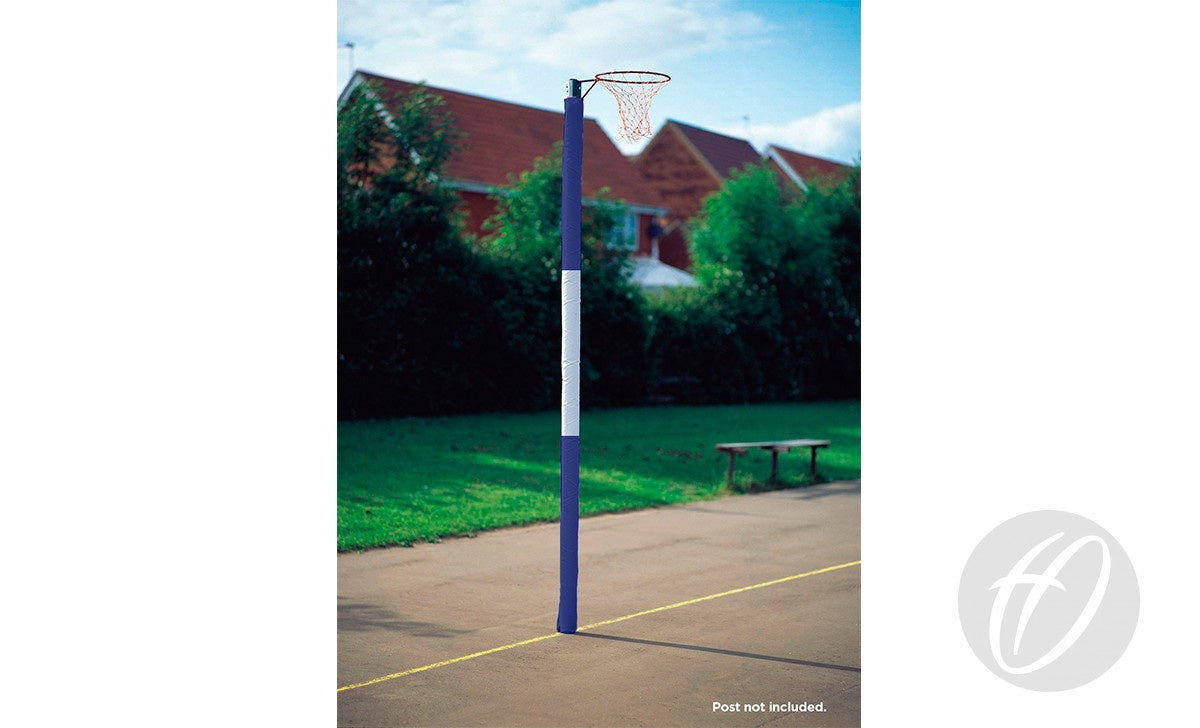 A netball hoop with a net is mounted on a blue post fitted with Harrod Sport Standard Netball Protectors - Three Colour, on an outdoor court. There's a bench nearby, trees behind, and red-roofed houses beyond the fence.