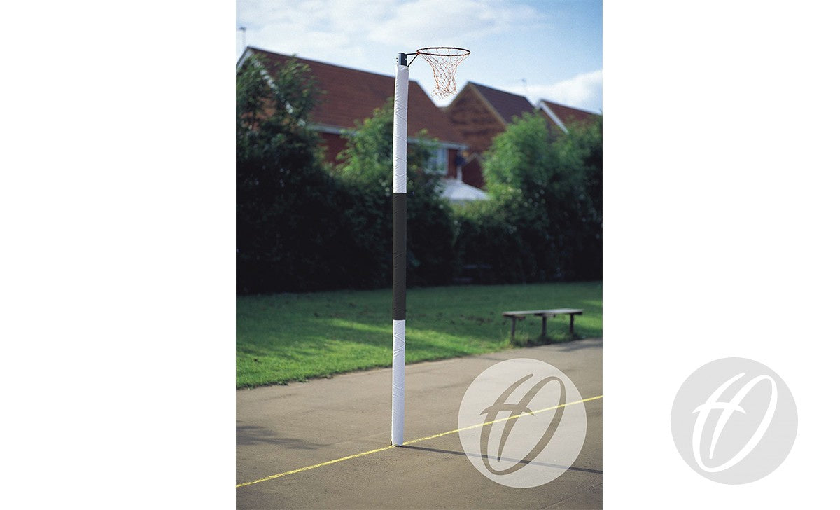 A Harrod Sport Standard Netball Protector - Three Colour covers the hoop and pole on an outdoor court marked by a yellow line. In the background are a bench, green foliage, and red-roofed houses beyond the trees.