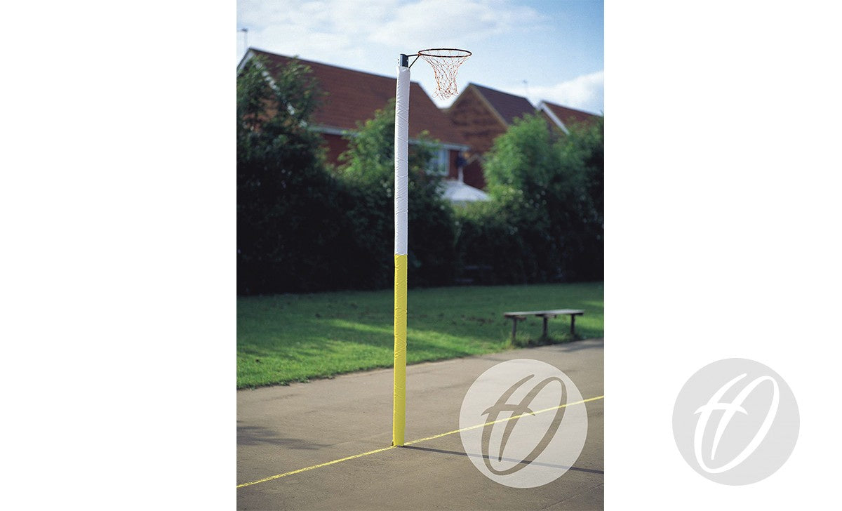 A Harrod Sport Standard Netball post with Two Colour Protectors stands on an outdoor court, featuring 50MM diameter posts. A bench and greenery sit in the background under a partly cloudy sky.