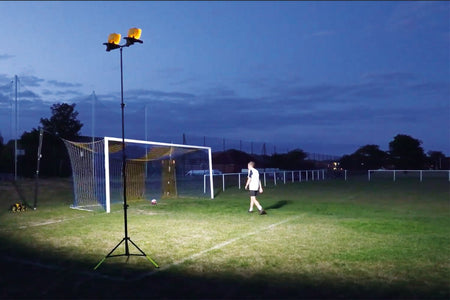 At dusk, a person stands near a goalpost on a soccer field, illuminated by the Nightsearcher Galaxy Pro Twin Portable Floodlights. A soccer ball lies on the grass as trees and fences fade into the dim background.