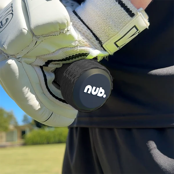 A close-up shows a person in white sports gloves holding the nub - Black cricket bat handle attachment by nub Sports, featuring its black silicone grip and logo. The scene is set outdoors on grass, with trees and a building in the background.