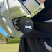 A close-up shows a person in white sports gloves holding the nub - Black cricket bat handle attachment by nub Sports, featuring its black silicone grip and logo. The scene is set outdoors on grass, with trees and a building in the background.