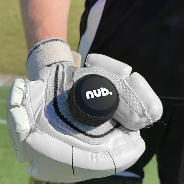 A person wearing a white cricket glove holds nub - Black by nub Sports, a silicone cricket grip for bat handles, with a green cricket field in the background.