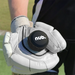 A person wearing a white cricket glove holds nub - Black by nub Sports, a silicone cricket grip for bat handles, with a green cricket field in the background.