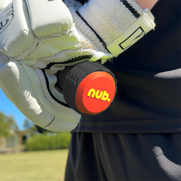A person wearing white sports gloves grips a cricket bat with a nub Sports silicone grip accessory, the nub – Orange Scorch, featuring a red and yellow logo on the end. The background shows green grass, trees, and a blurred building.