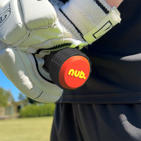 A person wearing white sports gloves grips a cricket bat with a nub Sports silicone grip accessory, the nub – Orange Scorch, featuring a red and yellow logo on the end. The background shows green grass, trees, and a blurred building.