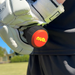 A person wearing white sports gloves grips a cricket bat with a nub Sports silicone grip accessory, the nub – Orange Scorch, featuring a red and yellow logo on the end. The background shows green grass, trees, and a blurred building.