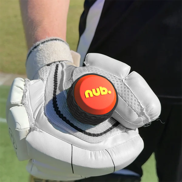 A person wearing a white cricket glove holds the "nub - Orange Scorch" by nub Sports, a black and orange silicone grip for cricket bats. A blurred outdoor sports field can be seen in the background.