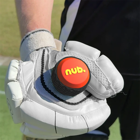 A person wearing a white cricket glove holds the "nub - Orange Scorch" by nub Sports, a black and orange silicone grip for cricket bats. A blurred outdoor sports field can be seen in the background.