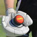 A person wearing a white cricket glove holds the "nub - Orange Scorch" by nub Sports, a black and orange silicone grip for cricket bats. A blurred outdoor sports field can be seen in the background.