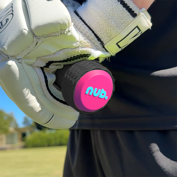 A close-up of a person wearing white sports gloves holding a cricket bat with the nub Sports "nub - Pink Six" pink and black silicone bat grip. A blurred grassy field and trees appear in the background.