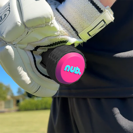 A close-up of a person wearing white sports gloves holding a cricket bat with the nub Sports "nub - Pink Six" pink and black silicone bat grip. A blurred grassy field and trees appear in the background.