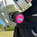 A close-up of a person wearing white sports gloves holding a cricket bat with the nub Sports "nub - Pink Six" pink and black silicone bat grip. A blurred grassy field and trees appear in the background.