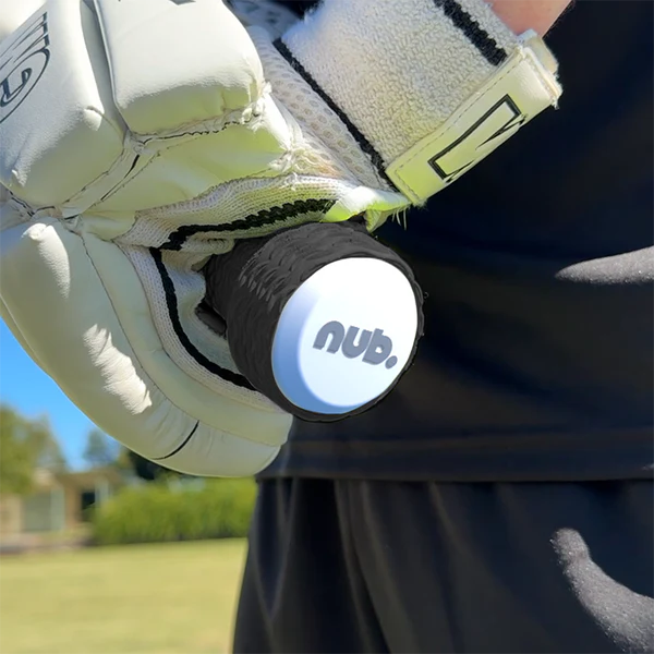 A close-up of a gloved hand holding a cricket bat handle with the nub - White by nub Sports; its white end cap and silicone grip stand out, while a green field and trees blur in the background.