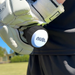 A close-up of a gloved hand holding a cricket bat handle with the nub - White by nub Sports; its white end cap and silicone grip stand out, while a green field and trees blur in the background.