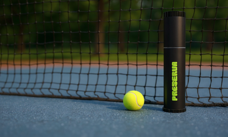A tennis ball sits on a blue court next to a black Preserva Rechargeable Tennis Ball Pressuriser canister, with the tennis net and green trees blurred in the background.
