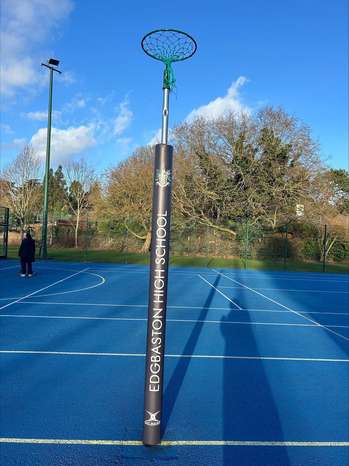 A pair of Gilbert Custom Printed Netball Post Protectors branded “Edgbaston High School” stands on a blue outdoor court under a clear sky, with trees and a fence in the background.