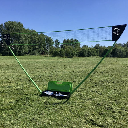 The Sure Shot Quick Fit 3 Junior Badminton Pack with OD shuttles by Sure Shot features green poles and a matching carrying case set up on grass, with rackets and shuttlecocks in the open case under a clear sky and trees in the background.