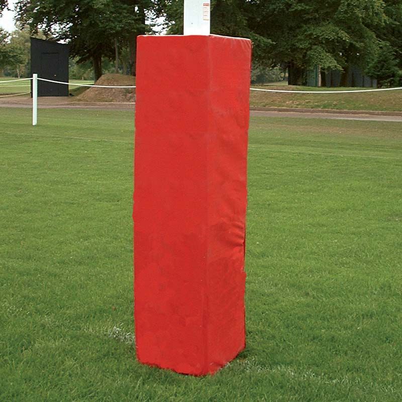 A Harrod Sport Millennium Rugby Post Protector, featuring red high-density foam padding, stands on a grassy field with trees and a rope barrier in the background.