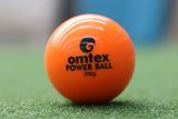 Close-up of an orange Omtex PowerBall, a 200g weighted cricket training ball by Omtex, placed on green artificial grass with a blurred blue and grey background.