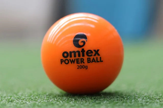 Close-up of an orange Omtex PowerBall, a 200g weighted cricket training ball by Omtex, placed on green artificial grass with a blurred blue and grey background.
