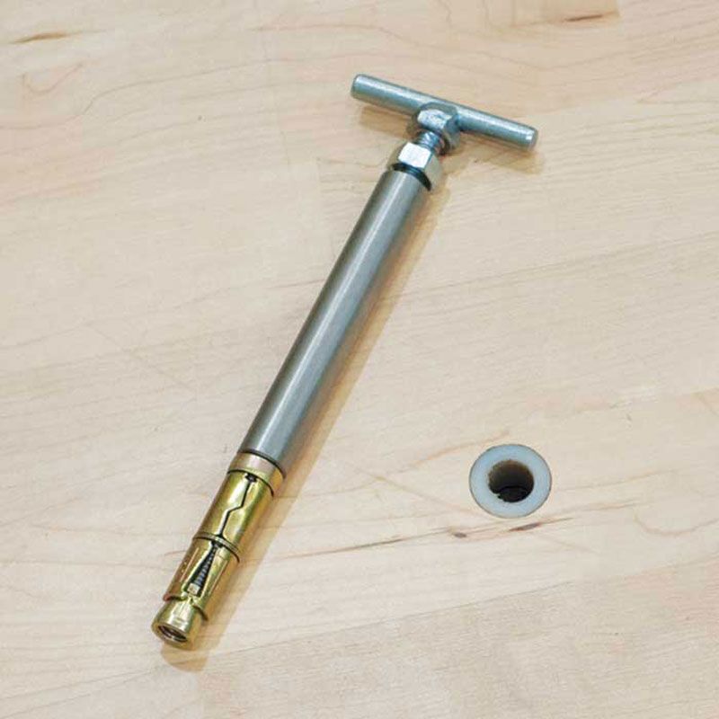 A Harrod Sport SH2 Sports Hall Floor Plug (210mm) installation tool with a T-handle rests on a wooden surface beside a circular opening, prepared for fitting the floor socket.