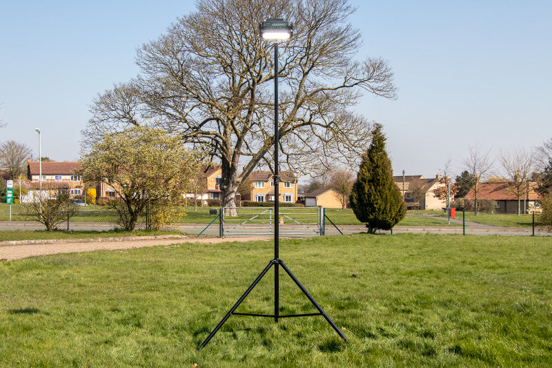 A Ritelite Sports Lite Portable Floodlight on a tall tripod stands in a grassy field with trees and houses in the background beneath a clear blue sky.