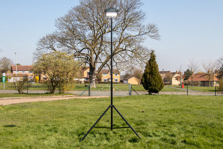 A Ritelite Sports Lite Portable Floodlight on a tall tripod stands in a grassy field with trees and houses in the background beneath a clear blue sky.