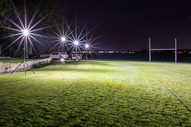 Ritelite Sports Lite Portable Floodlights brightly illuminate a rugby field at night, with goalposts visible and a white vehicle parked by a stone wall on the left.