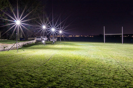 Ritelite Sports Lite Portable Floodlights brightly illuminate a rugby field at night, with goalposts visible and a white vehicle parked by a stone wall on the left.