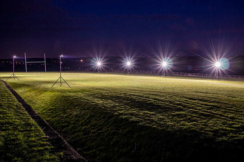 At night, the rugby field is brightly illuminated by Ritelite Sports Lite Portable Floodlights on tall stands, casting powerful light across the grass while the dark sky and distant scenery form the background.