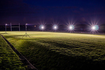 At night, the rugby field is brightly illuminated by Ritelite Sports Lite Portable Floodlights on tall stands, casting powerful light across the grass while the dark sky and distant scenery form the background.