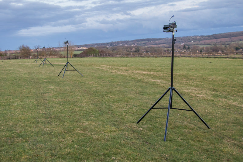 Three tripod-mounted cameras or sensors are set up in a grassy field under cloudy skies, accompanied by Ritelite Sports Lite Portable Floodlights, with hills and a village visible in the distance.