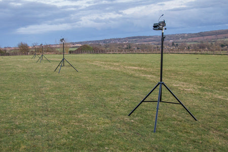 Three tripod-mounted cameras or sensors are set up in a grassy field under cloudy skies, accompanied by Ritelite Sports Lite Portable Floodlights, with hills and a village visible in the distance.