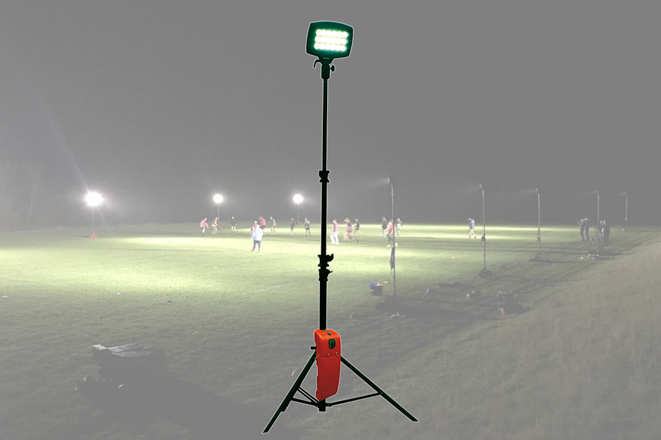 A Nightsearcher SportStar Portable LED Floodlight on a tripod brightly illuminates a night sports field with its powerful 20,000 lumen beam, while players compete nearby and mist drifts over the dimly lit, foggy background.