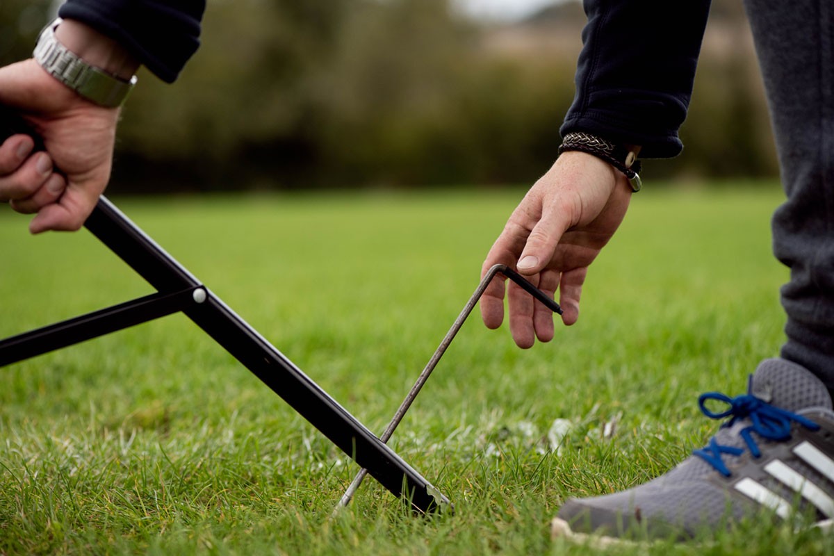 A person pegs a frame into grassy ground, with a Nightsearcher SportStar Portable LED Floodlight providing bright illumination nearby.