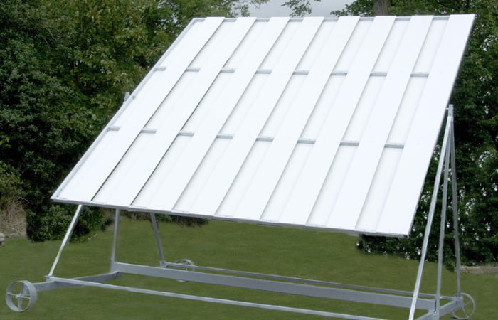 The Twenty/20 Sight Screen by Martin Berrill Sports is a large, white outdoor cricket sight screen with evenly spaced slats, a weather-resistant wheeled frame, and is set on grass with green trees behind it.