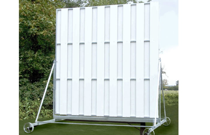 The Martin Berrill Sports Twenty/20 Sight Screen is a large, white rectangular screen with vertical panels and a weather-resistant wheeled frame, positioned outdoors on grass near trees under an open sky.