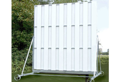 The Martin Berrill Sports Twenty/20 Sight Screen is a large, white rectangular screen with vertical panels and a weather-resistant wheeled frame, positioned outdoors on grass near trees under an open sky.