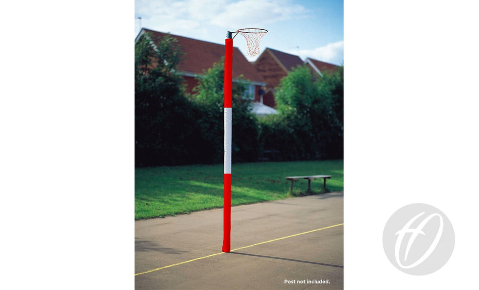 A Harrod Sport Standard Netball Post with Three Colour foam-filled, vinyl-coated nylon protectors stands on an outdoor court. Grass, trees, houses, a bench near the net, and the yellow boundary line are visible in the background.