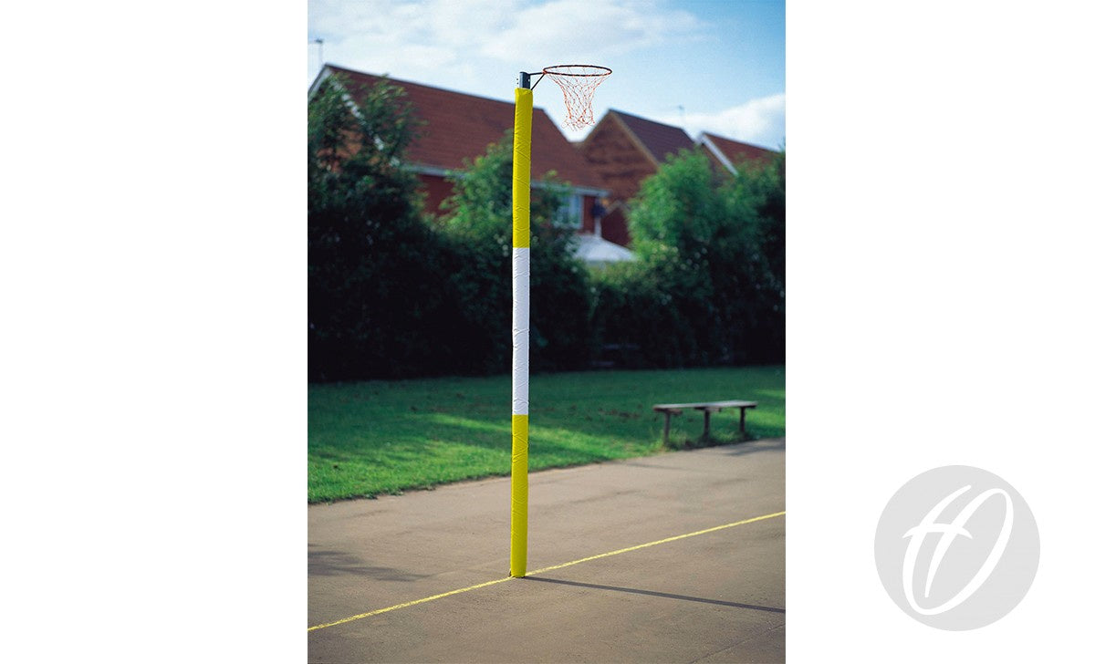 A yellow netball post with a hoop and net features Harrod Sport Standard Netball Protectors - Three Colour with foam-filled pads, standing on an outdoor court surrounded by grass, trees, benches, and red-roofed houses under a blue sky.