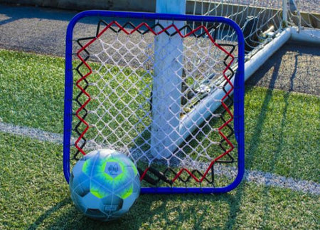 A Samba Sports Large Handheld Re-bounder with a blue frame and red border sits on artificial turf near a goalpost, with a soccer ball resting against it—ideal for goalkeeping coaching sessions.
