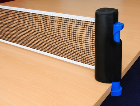 A close-up of a Sure Shot Matthew Syed Flex Net with black posts and blue clasps attached to the edge of a light wooden table. The Sure Shot net is tightly stretched across the table, ready for play.