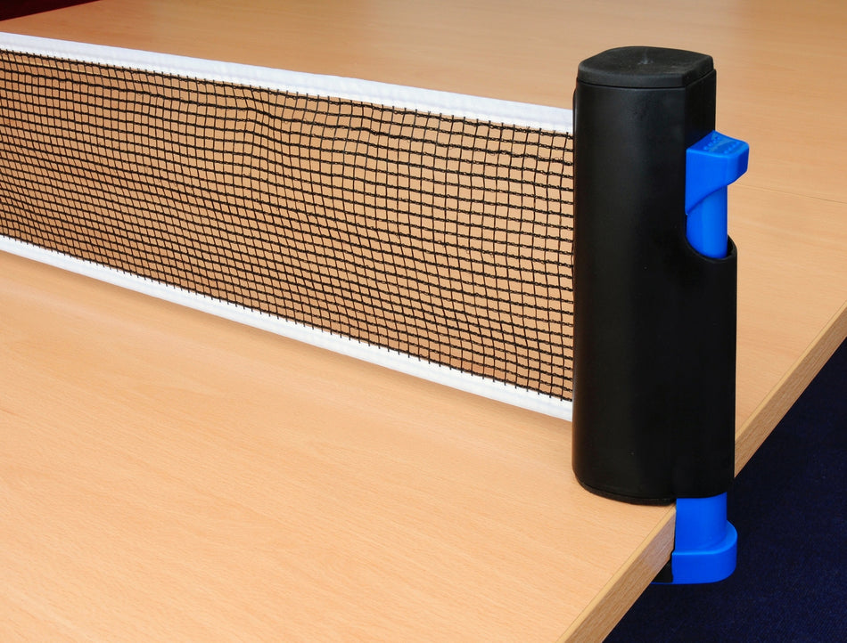 A close-up of a Sure Shot Matthew Syed Flex Net with black posts and blue clasps attached to the edge of a light wooden table. The Sure Shot net is tightly stretched across the table, ready for play.