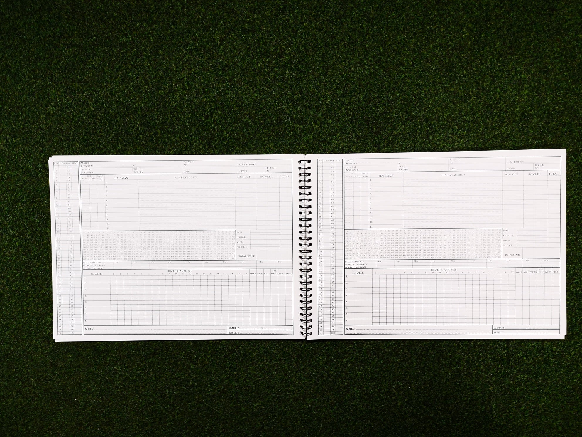 A Gray Nicolls 112 Innings Scorebook by Gray Nicolls, spiral-bound and open to blank pages filled with tables and charts, rests on green grass—ready for scoring your next cricket match.