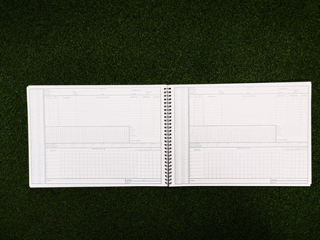 A Gray Nicolls 112 Innings Scorebook by Gray Nicolls, spiral-bound and open to blank pages filled with tables and charts, rests on green grass—ready for scoring your next cricket match.