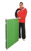 A man in a red and black jacket stands beside the Butterfly Junior Compact Table Tennis Table by Butterfly, shown folded against a white background to highlight its space-saving design.