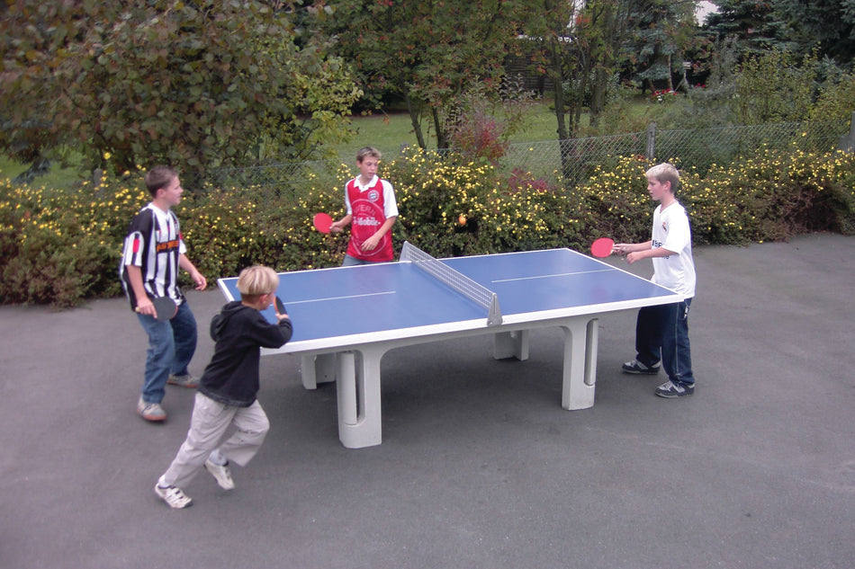 Four boys play doubles on the Butterfly Park Polymer Concrete 45SQ Table Tennis Table by Butterfly—a durable, blue outdoor table set amid greenery and yellow flowers, designed for permanent outdoor use. All are focused and holding paddles.