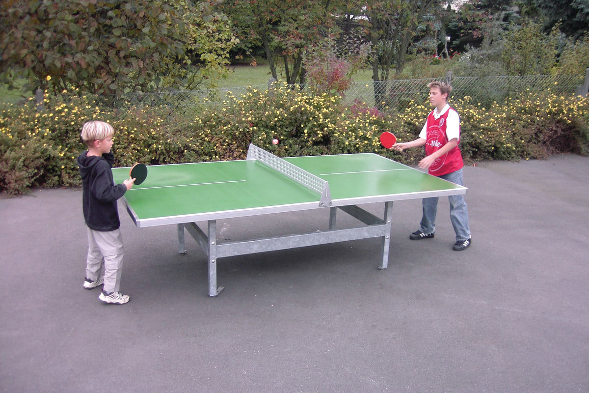 Two boys play table tennis on a blue Butterfly S2000 Polymer Concrete/Steel table with square corners. Surrounded by trees, both hold paddles and appear focused. The sturdy Butterfly table is designed for permanent outdoor use.