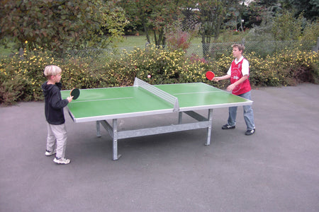 Two boys play table tennis on a blue Butterfly S2000 Polymer Concrete/Steel table with square corners. Surrounded by trees, both hold paddles and appear focused. The sturdy Butterfly table is designed for permanent outdoor use.